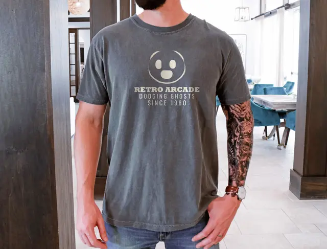 Person wearing a Pepper tee printed with RETRO ARCADE, DODGING GHOSTS SINCE 1980