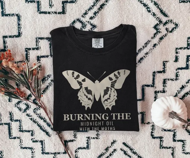 Person wearing a Black tee printed with BURNING THE, MIDNIGHT OIL, WITH THE MOTHS